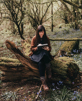 Natalie Toms sitting on a moss-covered tree stump. She is holding a sketchbook and looking down at what she is drawing. She is Surrounded by trees and bluebells in spring. Natalie wears a brown jacket, a grey dress, and brown boots. Photo by Rebecca Rees.